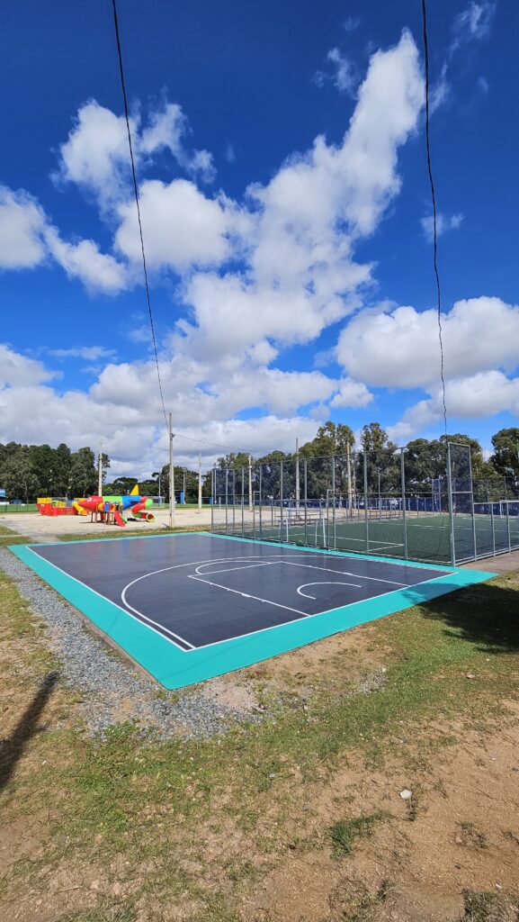 Uma fotografia ao ar livre de ângulo médio em um dia claro, mostrando um complexo esportivo comunitário. No primeiro plano, uma quadra de basquete de asfalto preto limpo com linhas brancas nítidas é cercada por um perímetro turquesa brilhante. À direita dela, uma quadra de futsal maior é cercada por uma alta cerca de arame galvanizado e possui uma superfície de grama sintética verde com postes de gol. Ao longe, à esquerda, há um parquinho infantil colorido em forma de avião sobre areia, cercado por grama verde. Vários postes de luz estão espalhados pelo local. Ao fundo, uma densa linha de árvores verdes escuras se estende. O céu azul brilhante está preenchido com muitas nuvens brancas e macias. Duas linhas de energia preta cruzam diagonalmente o céu do canto superior esquerdo para o canto superior direito.