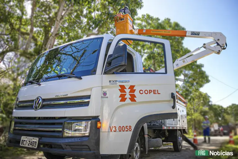 Um caminhão de manutenção da Copel (Volkswagen Delivery Express), de cor branca, com a porta do motorista aberta. Na porta, estão os logotipos da Copel e do Governo do Paraná, e o número do veículo V3 0058. No alto, um técnico está dentro de um cesto elevatório de cor laranja acoplado ao braço hidráulico, realizando um trabalho perto de cabos elétricos entre árvores. Um segundo técnico está no solo, ao fundo, ao lado de cones de sinalização.