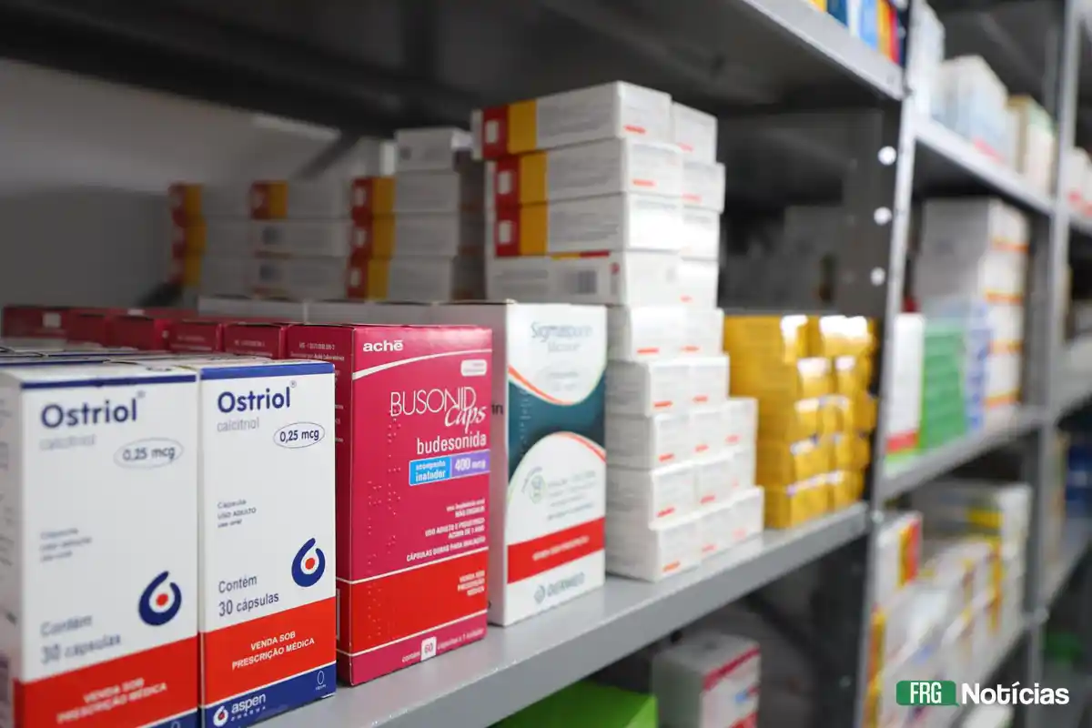 Prateleiras de metal cinza organizadas com diversas caixas de medicamentos em uma farmácia. Em destaque, caixas de Ostriol e Busonid. No canto inferior direito, a logo FRG Notícias.