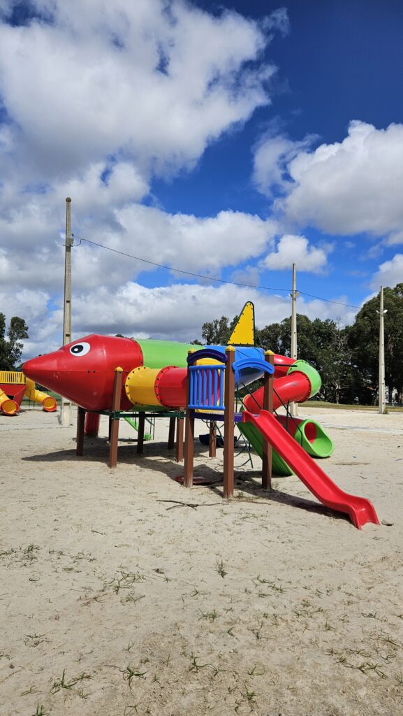 Foto vertical de um parquinho em formato de avião estilizado, instalado sobre um banco de areia. A estrutura é composta por tubos coloridos em vermelho, verde, amarelo e azul. Na frente, a cabine vermelha tem o desenho de um olho. O brinquedo possui diversos escorregadores, incluindo um reto na cor vermelha e outros em espiral. O cenário ao fundo apresenta árvores altas, postes de iluminação e um céu azul intenso com nuvens brancas e densas.
