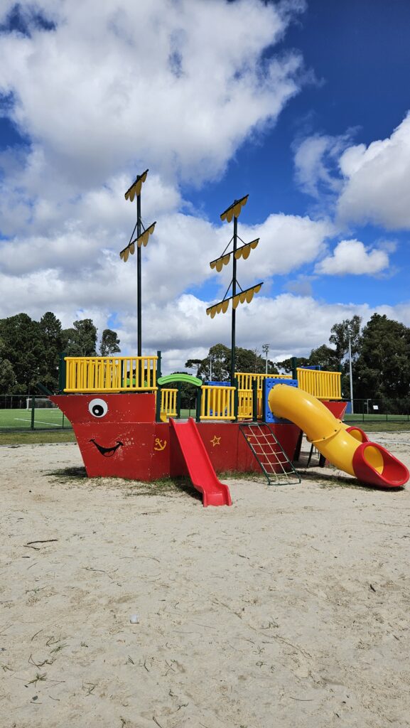 Fotografia vertical de um playground colorido em formato de navio pirata, posicionado sobre uma grande área de areia clara. O casco do navio é vermelho com um rosto sorridente desenhado na proa. Possui dois mastros pretos com velas amarelas estilizadas, um escorregador reto vermelho e um escorregador em tubo caracol amarelo. Ao fundo, árvores verdes e um céu azul vibrante com nuvens brancas volumosas.