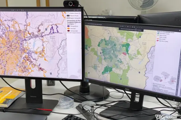 Dois monitores de computador exibindo um Sistema de Informações Geográficas (SIG). O monitor à esquerda mostra mapas de transporte público com linhas coloridas, enquanto o monitor à direita exibe um mapa de calor sobre o Índice de Vulnerabilidade Social na Região Metropolitana de Curitiba.