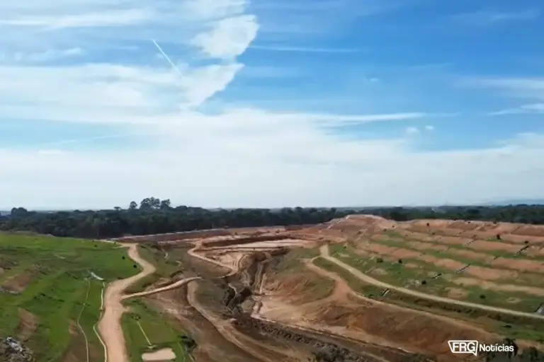 Uma vista aérea de um grande aterro sanitário em camadas. Do lado esquerdo e no topo, vê-se grama verde e árvores. Do lado direito e no centro, a montanha de lixo e a terra revirada formam degraus e valetas de terra marrom. Uma estrada de terra serpenteia pelo local. O céu é azul claro com nuvens brancas alongadas. No canto inferior direito, há uma pequena marca d'água branca que diz FRG Notícias.