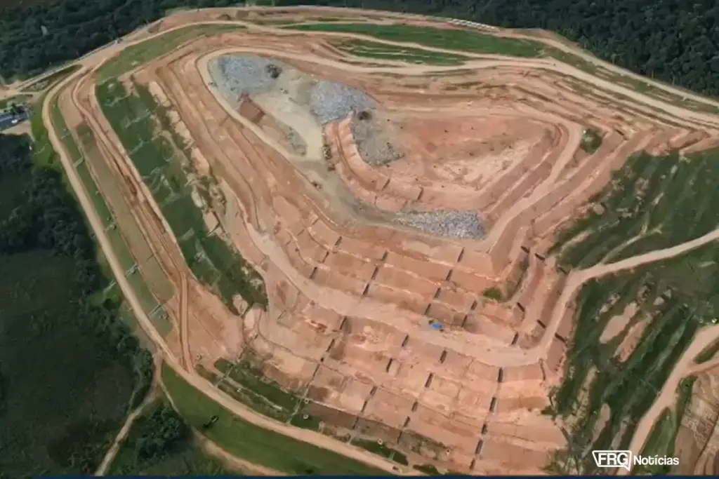 Uma vista aérea ortogonal e mais próxima de uma nova seção monumental e vazia de um aterro sanitário. O terreno foi escavado em cortes e degraus de engenharia precisos e simétricos em solo marrom-avermelhado e creme, sem lixo visível. O padrão é geométrico e organizado. No fundo e nas laterais, a densa floresta escura contrasta fortemente com a terra nua. O céu não é visível. No canto inferior direito, a marca d'água FRG Notícias.