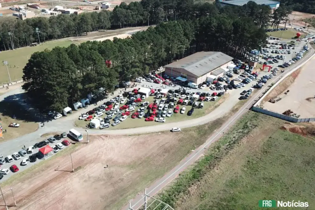 Vista aérea do Centro MultiEventos em Fazenda Rio Grande durante um evento automotivo. O gramado e o pátio ao redor de um grande galpão estão repletos de carros estacionados e pequenas tendas brancas. Há uma área de mata preservada ao fundo e um campo de futebol à esquerda.