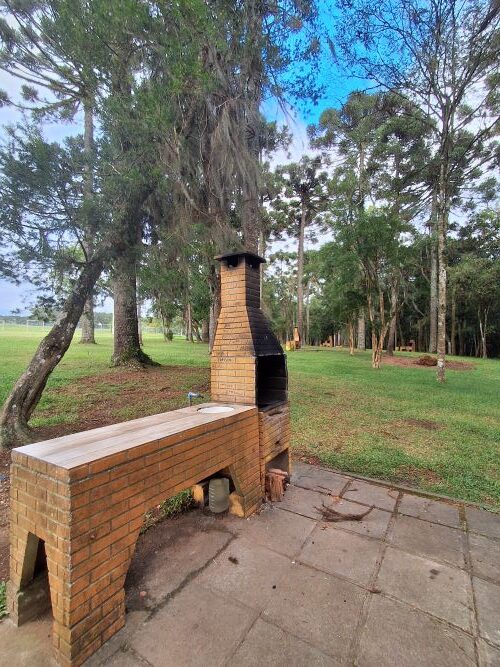 Uma churrasqueira de tijolos ao ar livre, com pia e bancada acopladas, situada em uma área de lazer pavimentada com blocos de concreto. O fundo é composto por um amplo gramado verde e diversas araucárias sob um céu claro.
