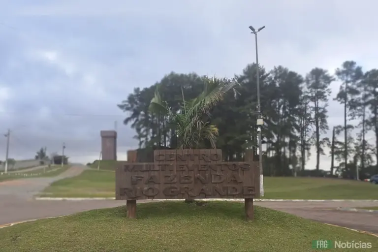 Uma placa de madeira rústica e esculpida, com o texto "CENTRO MULTIEVENTOS FAZENDA RIO GRANDE", está fixada em um canteiro circular de grama na entrada do local. Ao fundo, uma alameda de pinheiros altos sob um céu nublado. À direita, um poste de iluminação alto e, ao fundo à esquerda, uma torre de observação.