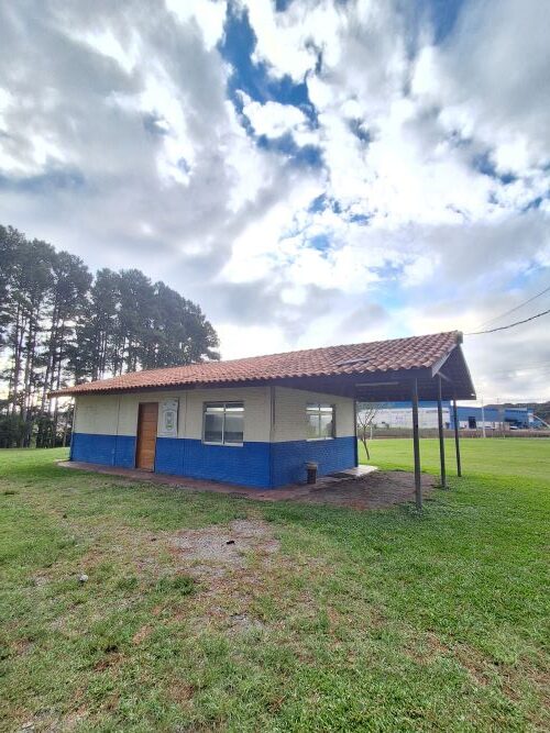 Uma construção de alvenaria térrea com telhado de telhas cerâmicas e paredes pintadas de branco e azul na base. O edifício está situado em um gramado verde, com uma fileira de pinheiros altos à esquerda e um galpão industrial ao fundo, sob um céu com nuvens carregadas e algumas aberturas azuis.