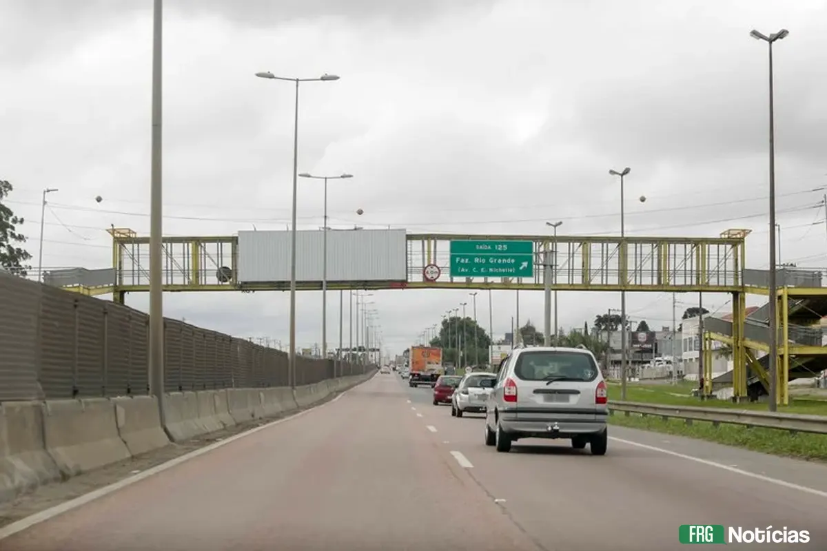 Fotografia ao nível da estrada na BR-116 em Fazenda Rio Grande, mostrando uma passarela metálica amarela sobre a rodovia. Veículos trafegam sob a estrutura em um dia nublado. Ao fundo, placas de sinalização indicam a saída 125. No canto inferior direito, o logotipo FRG Notícias"