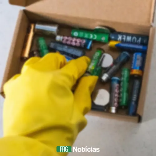 Mão com luva amarela pegando uma pilha de dentro de uma caixa de papelão cheia de baterias e pilhas diversas. Abaixo, a marca d'água FRG Notícias.