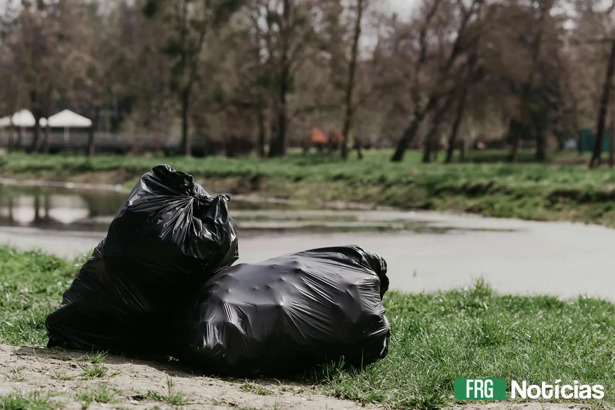 A imagem mostra dois grandes sacos de lixo pretos e cheios deitados sobre a grama verde às margens de um rio desfocado. Ao fundo, árvores e vegetação esparsa. No canto inferior direito, há o logotipo FRG Notícias.
