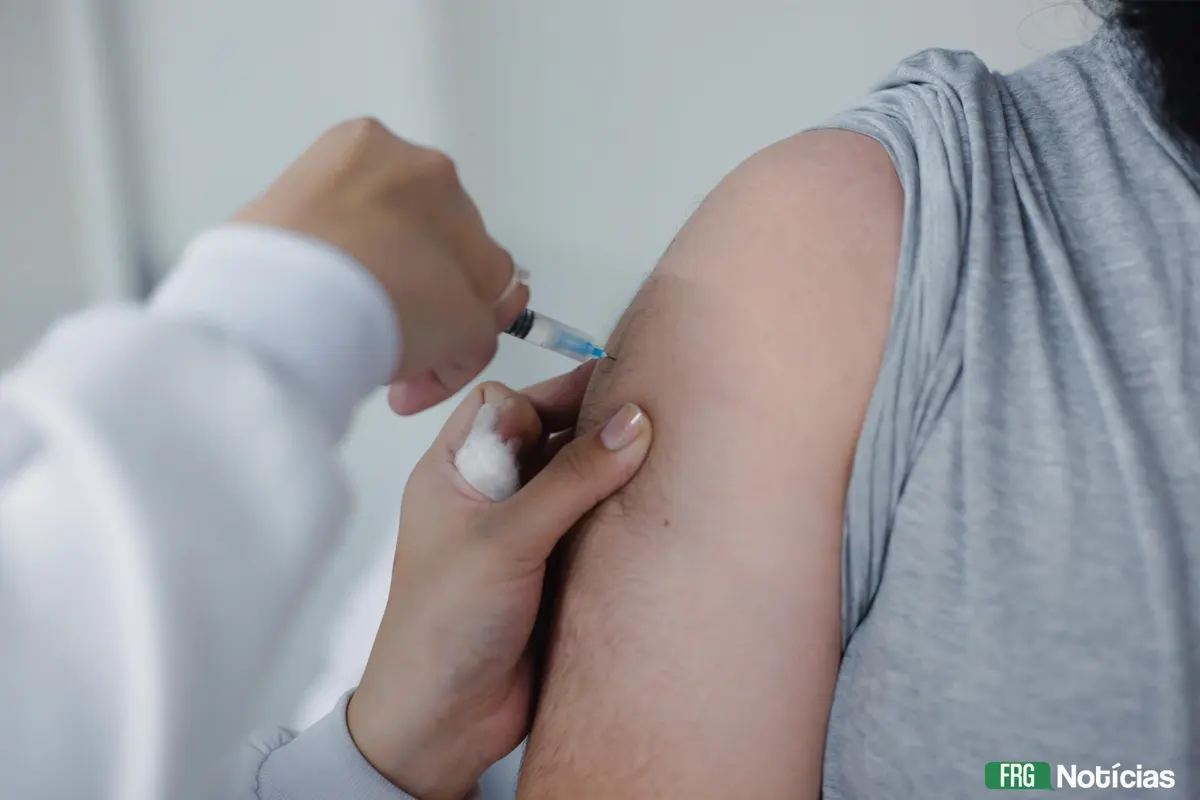 Close-up do braço de uma pessoa recebendo uma vacina. Uma profissional de saúde, vestindo jaleco branco, segura uma seringa e aplica o imunizante no ombro do paciente. A profissional utiliza um pedaço de algodão para apoiar a aplicação. No canto inferior direito, o logotipo verde e branco do portal FRG Notícias.