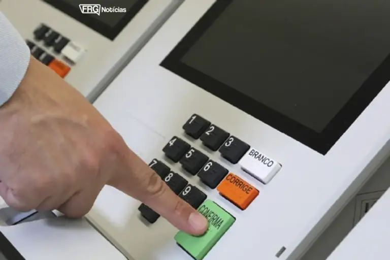 Close-up da mão de um eleitor pressionando o botão verde CONFIRMA no teclado de uma urna eletrônica brasileira. O logo do FRG Notícias está visível no canto superior.
