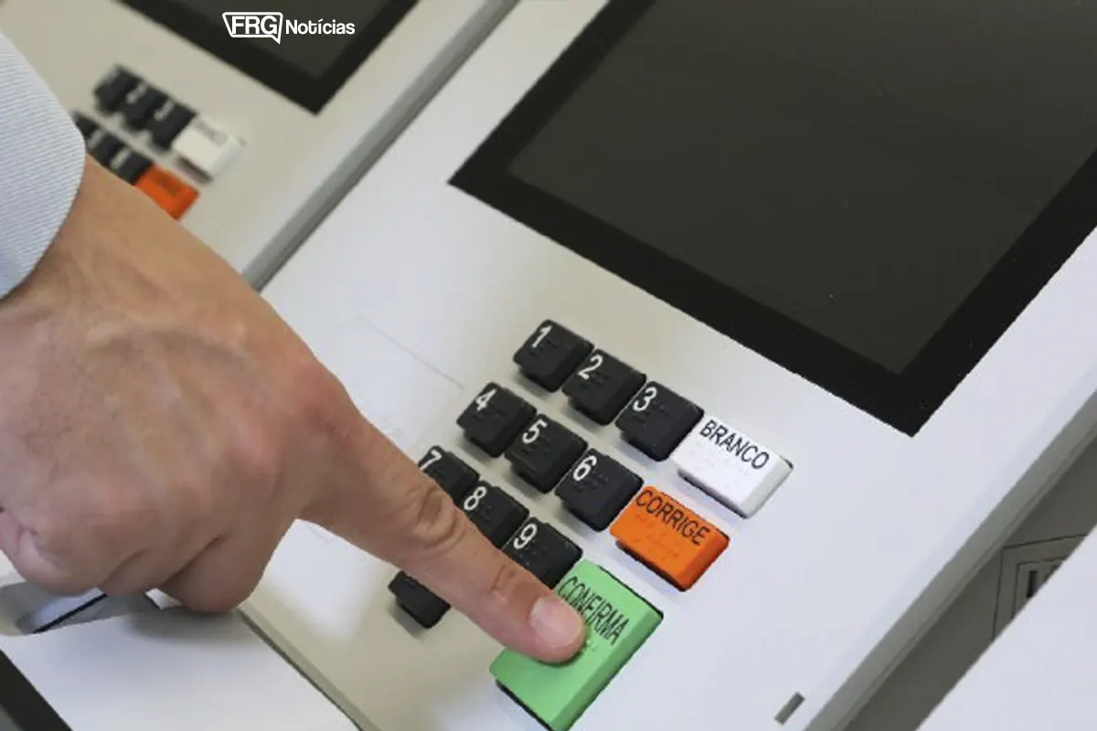 Close-up da mão de um eleitor pressionando o botão verde CONFIRMA no teclado de uma urna eletrônica brasileira. O logo do FRG Notícias está visível no canto superior.