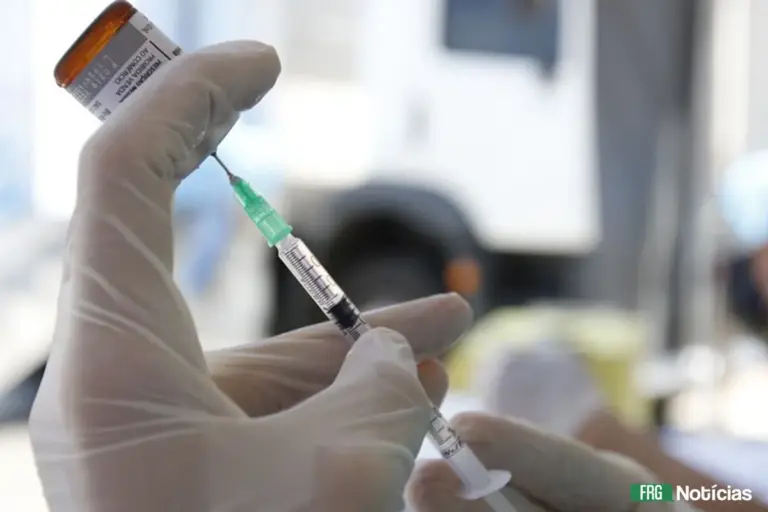 Profissional de saúde usando luvas brancas segura uma seringa e um frasco de vacina, preparando a dose para aplicação. No canto inferior direito, há o logotipo FRG Notícias