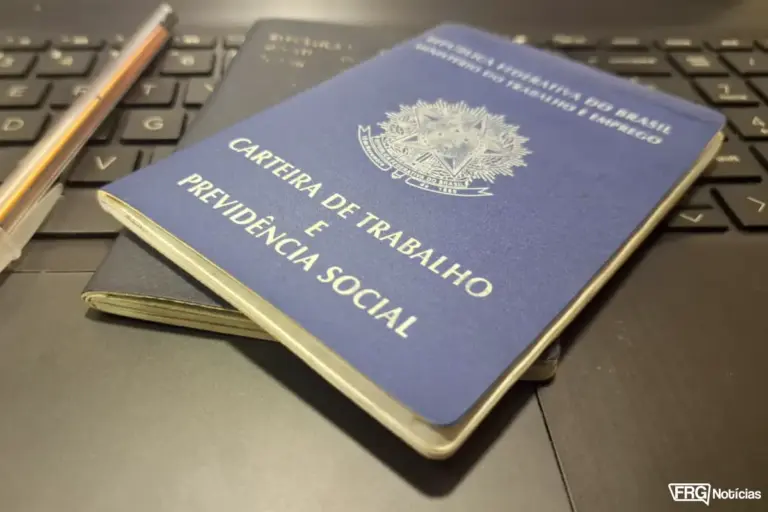 Foto em close de uma Carteira de Trabalho e Previdência Social (CTPS) física de cor azul sobreposta a outra carteira idêntica, posicionadas em cima do teclado preto de um notebook. No canto superior esquerdo aparece parte de uma caneta esferográfica transparente. No canto inferior direito, consta o logotipo oficial do portal FRG Notícias.