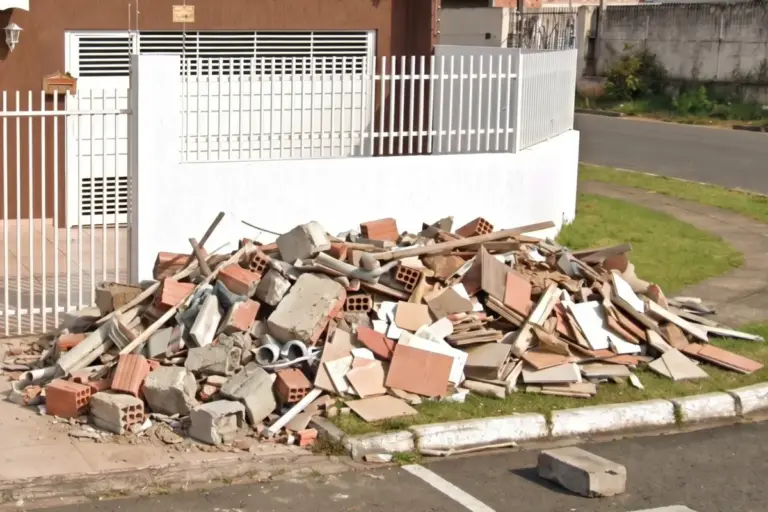 Pilha de entulho de construção acumulada na calçada em frente a uma residência, contendo restos de tijolos, azulejos quebrados, concreto e pedaços de madeira.