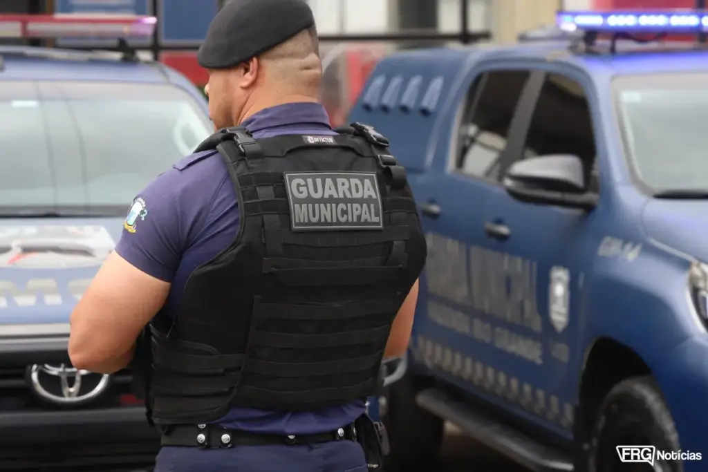 Agente da Guarda Municipal de Fazenda Rio Grande de costas, utilizando colete balístico com a identificação Guarda Municipal. Ao fundo, viaturas policiais azuis com giroflex ligados, simbolizando o policiamento ostensivo e a segurança pública no município.