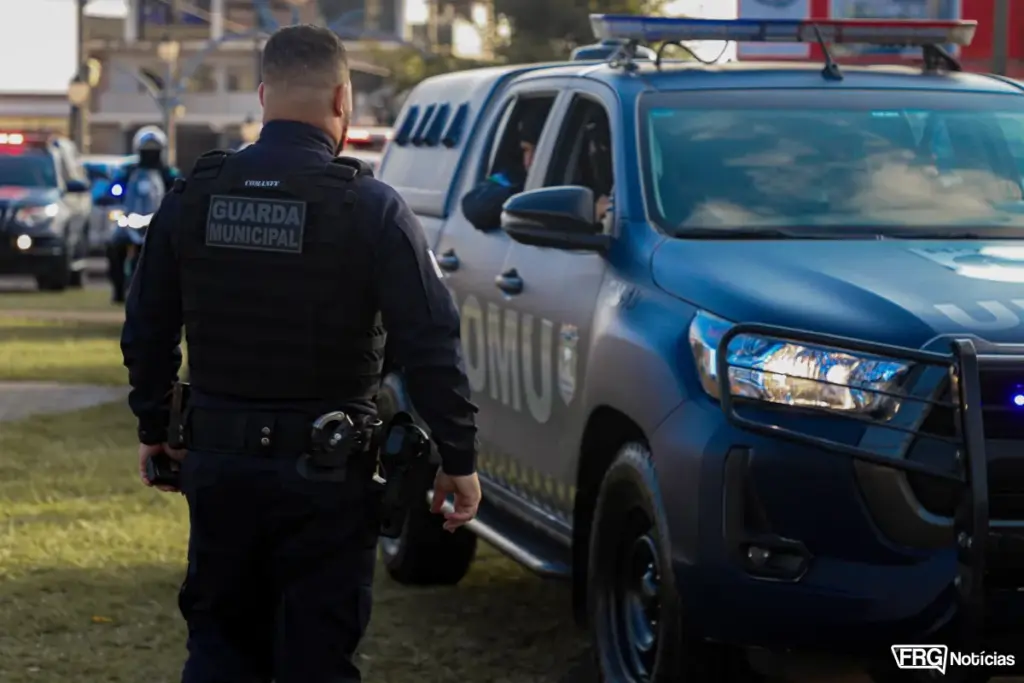 Agente da Guarda Municipal de Fazenda Rio Grande em primeiro plano, equipado com colete tático e rádio comunicador. Ao lado, uma viatura da ROMU com faróis acesos em operação de patrulhamento urbano. No fundo, é possível ver outras viaturas e motocicletas da corporação em uma área gramada durante o entardecer.