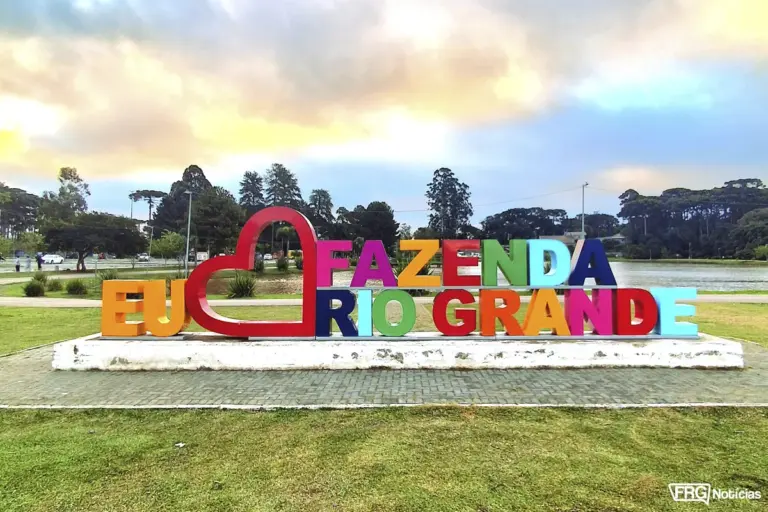 Letreiro colorido com a frase "Eu Amo Fazenda Rio Grande" em destaque no Centro Esportivo Ayrton Senna, antigo Parque Verde, com o lago e araucárias ao fundo sob um céu de entardecer em Fazenda Rio Grande, PR.