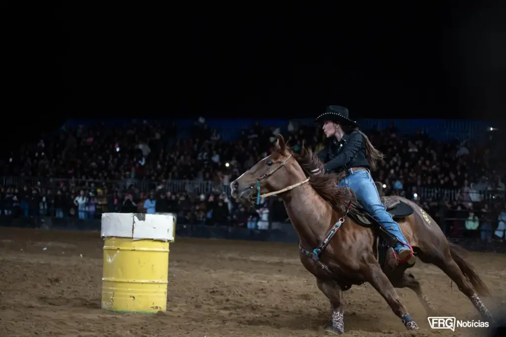 Amazona e cavalo quarto de milha em alta velocidade contornando tambor amarelo na Prova dos Três Tambores da Expo Fazenda, em Fazenda Rio Grande. Arena lotada ao fundo à noite.
