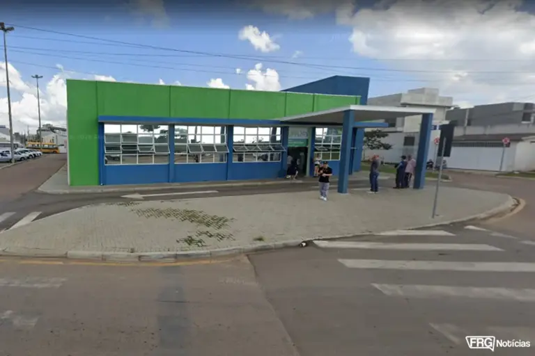 Uma imagem de rua em perspectiva ao nível dos olhos da fachada da UPA 24h, uma clínica médica de emergência em Fazenda Rio Grande, Paraná. O edifício moderno apresenta paredes verdes e azuis brilhantes com grandes janelas horizontais. Um pórtico coberto de azul estende-se sobre a entrada principal, apoiado por colunas, e um letreiro sutil UPA 24h é visível. Várias pessoas, em trajes casuais de rua, estão em pé no pavimento em frente à entrada. O pavimento é pavimentado e apresenta uma faixa de pedestres proeminente em primeiro plano, com linhas amarelas e brancas pintadas. O céu é azul claro com nuvens brancas esparsas. Postes de iluminação de rua altos estão localizados à esquerda e à direita. Uma rua lateral estende-se para a esquerda, com carros estacionados visíveis ao longe. A arquitetura envolvente é predominantemente residencial e comercial de baixa densidade. A perspectiva da câmera captura a profundidade e a escala do edifício e do seu ambiente urbano. O selo Google é visível na parte superior central. Uma marca de água FRG Notícias está localizada no canto inferior direito.