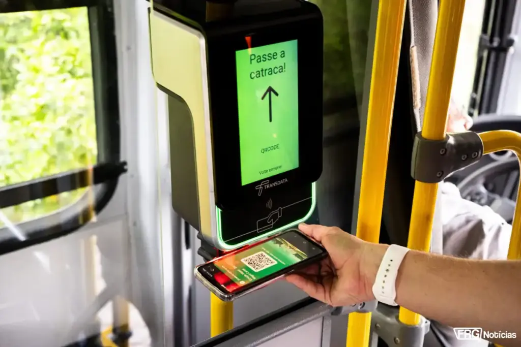 Mão de passageiro aproximando um smartphone com QR Code de um validador de passagens eletrônico da Transdata dentro de um ônibus. A tela do dispositivo exibe a mensagem 'Passe a catraca!' em fundo verde. No canto inferior direito, o logotipo FRG Notícias.