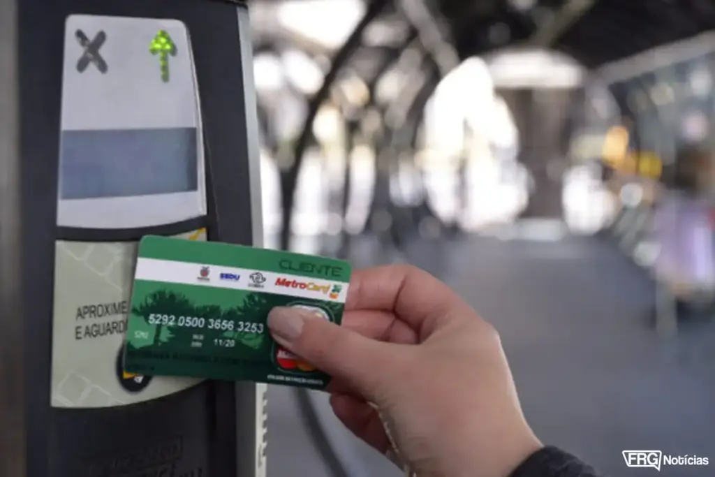 Close de uma mão feminina aproximando o cartão Metrocard (modelo verde, categoria Cliente) de um validador de passagens eletrônico em uma estação tubo ou terminal. O validador exibe uma seta verde iluminada e a instrução 'Aproxime e aguarde'. No canto inferior direito, o logotipo do FRG Notícias.