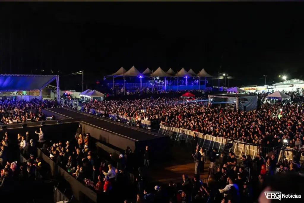 Visão panorâmica noturna da arena de shows na Expo Fazenda, em Fazenda Rio Grande, mostrando uma grande multidão, passarelas de palco, áreas VIP com tendas iluminadas e a organização do evento.