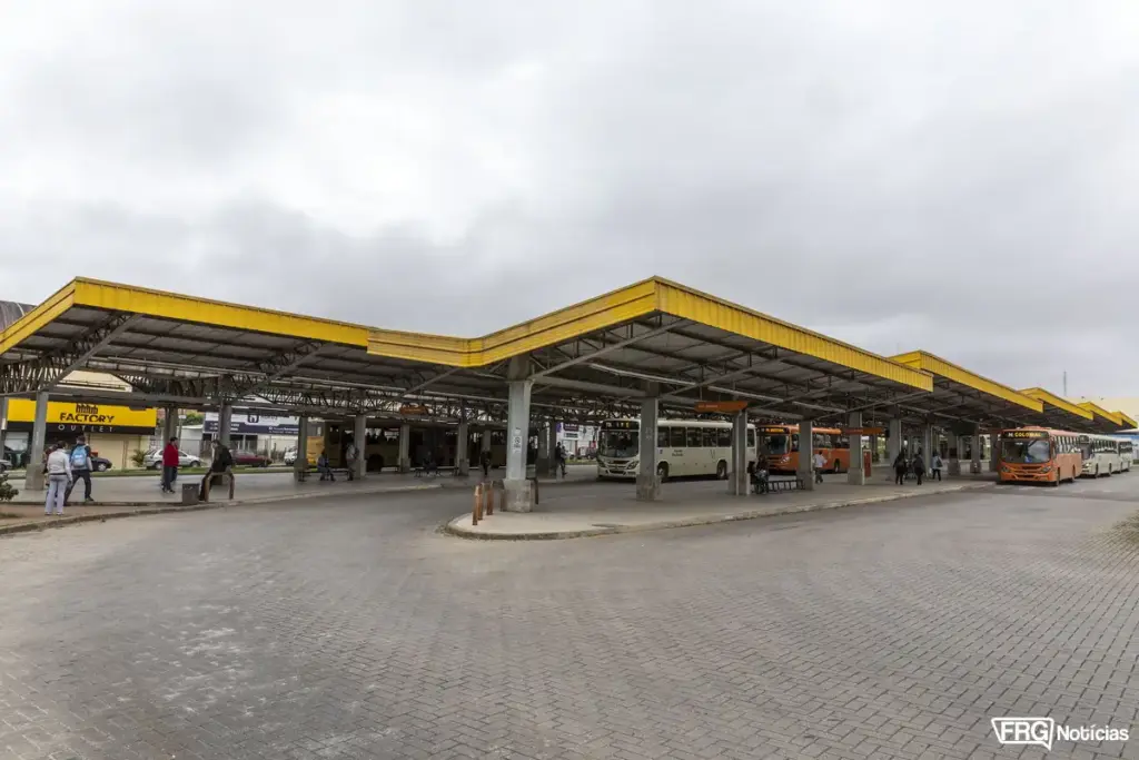 Vista ampla do Terminal de Integração de Fazenda Rio Grande sob um céu nublado. A imagem mostra a estrutura metálica de cobertura amarela, diversas plataformas de embarque com ônibus das linhas Ipê e Colonial estacionados, e passageiros aguardando ou caminhando. Ao fundo, é visível o comércio local, incluindo a fachada da loja Factory Outlet. No canto inferior direito, o logotipo do FRG Notícias.